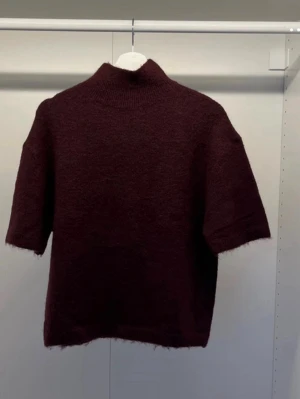 Mörk vinröd stickad topp - Mysig mörk vinröd stickad topp med korta ärmar och hög krage. Perfekt för lager på lager och ger en avslappnad men ändå stilren look. Passar till både jeans och kjol.
