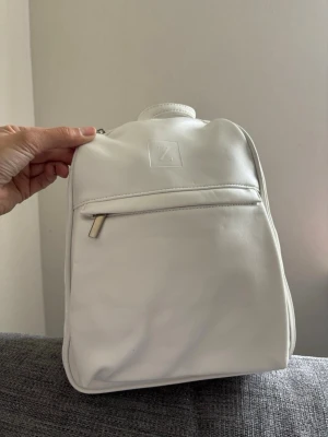 Vit minimalistisk ryggsäck i skinnimitation - Snygg vit ryggsäck i skinnimitation med stilren design och präglad Z-logga framtill. Väskan har en yttre ficka med dragkedja och diskreta sömmar. Perfekt för dig som gillar enkel och modern stil. Mått 30x22x11 cm 