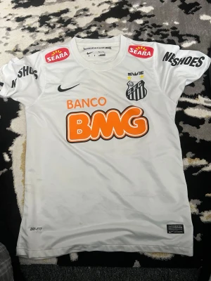 Santos FC Neymar Jr matchtröja - Vit Santos FC matchtröja från Nike med Neymar Jr och nummer 11 på ryggen. Tröjan har flera sponsorloggor, klubbmärke på bröstet och är tillverkad i ett lätt, ventilerande material. Perfekt för dig som älskar brasiliansk fotboll och vill sticka ut på planen.