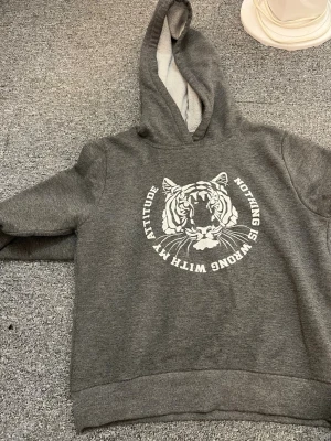 Grå hoodie med tigertryck - Snygg grå hoodie med huva och ett stort vitt tigertryck på bröstet. Texten runt motivet lyder 'Nothing is wrong with my attitude'. Hoodien har mjukt material och ribbade muddar vid ärmar och nederkant. Perfekt för dig som gillar streetstyle och djurmotiv.