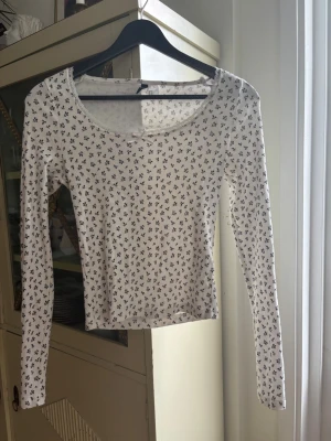 Vit långärmad topp med blommönster - Söt vit långärmad topp med små svartblå blommor över hela tyget. Modellen har en rundad, lite djupare urringning och en liten rosett framtill. Tunn och mjuk bomullskänsla, perfekt till jeans eller kjol.