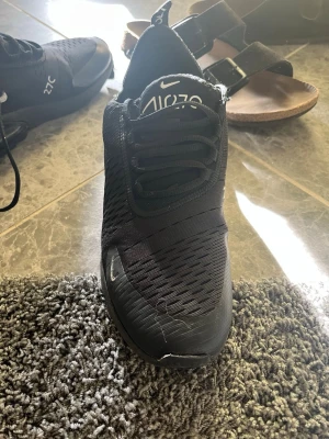 Svarta Nike Air Max 270 sneakers - Snygga svarta Nike Air Max 270 sneakers med vit logga och textdetaljer. Skorna har en luftig mesh-överdel, synlig Air-enhet i hälen och klassisk snörning. Perfekta för dig som gillar sportig och modern stil.