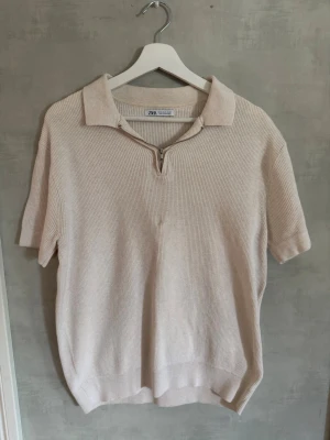 Beige pikétröja från Zara - Säljer en beige pikétröja från Zara med kort ärm och krage. Tröjan har en liten dragkedja vid halsen och är gjord i ett mjukt, ribbat material. Perfekt för en avslappnad men ändå stilren look. Skick 8/10, Storlek M, hör av dig vid frågor🙌
