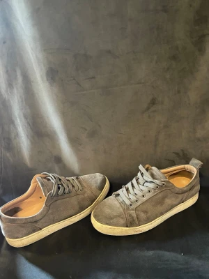 Louboutin  - Snygga sneakers i grå mocka med klassisk låg profil och vita sulor. Skorna har röda yttersulor som ger en cool kontrast och stilren look. Perfekta för dig som gillar en enkel men ändå unik stil. Skosnören i matchande grå färg.