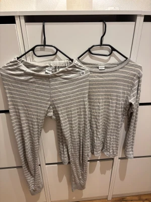 Randig pyjamas Polarn O. Pyret 146/152 - Tvådelad pyjamas från Polarn O. Pyret i grått och vitt med tunna horisontella ränder. Setet består av långärmad tröja och byxor med resår i midjan. Tillverkad i mjuk ekologisk bomull, perfekt för sköna nätter hemma.