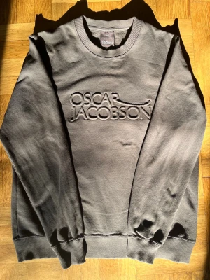 Oliv grön sweatshirt från Oscar Jacobson - Snygg grå sweatshirt från Oscar Jacobson med upphöjt logotyptryck på bröstet. Tröjan har rund halsringning, ribbade muddar vid ärmslut och nederkant samt en avslappnad passform. Perfekt för en stilren och enkel look.