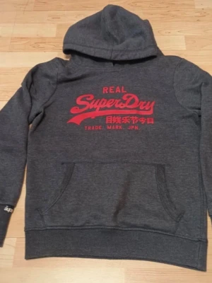 Grå Superdry hoodie med röd logga - Mörkgrå hoodie från Superdry med stor röd logga och japansk text på bröstet. Klassisk känguruficka framtill och huva. Perfekt för en avslappnad och cool stil.