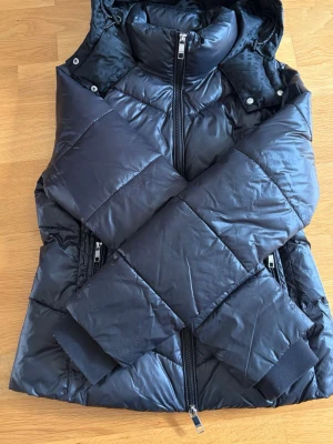 Svart pufferjacka med huva från Hugo Boss  - Snygg svart pufferjacka med avtagbar huva och hög krage. Jackan har två dragkedjeförsedda fickor fram, ribbade muddar vid ärmsluten och quiltad design. Perfekt för kalla vinterdagar och riktigt varm. Ser mörkblå ut men den är svart 