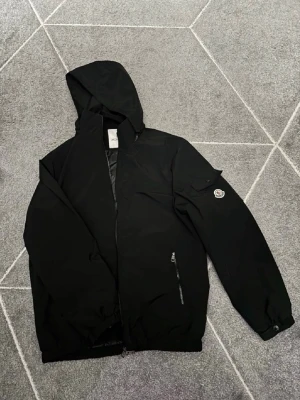 Svart vindjacka från Moncler - Snygg svart vindjacka från Moncler med huva och dragkedja framtill, dragkedjan är trasig men man kan fixa de!Jackan har en ficka med lock och Moncler-logga på ärmen samt två dragkedjefickor. Perfekt för blåsiga dagar och enkel att matcha med allt. 