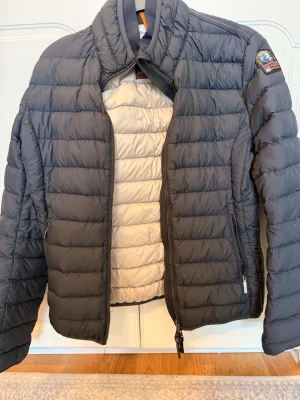 Mörkgrå dunjacka från Parajumpers - Snygg mörkgrå dunjacka från Parajumpers med quiltad design och hög krage. Jackan har dragkedja framtill och en patch med logga på ärmen. Insidan är ljusare och jackan har en modern, sportig look.