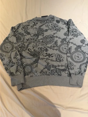 Grå sweatshirt Versace Jeans Couture - Snygg grå sweatshirt från Versace Jeans Couture med svart barockinspirerat mönster och logodetaljer. Tröjan har rund hals, ribbade muddar vid ärmslut och nederkant samt en avslappnad passform. Perfekt för dig som gillar statement-plagg.