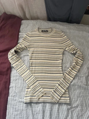 Randig ribbad topp från Gina Tricot - Säljer en långärmad, ribbad topp från Gina Tricot i beige, grått och vitt. Toppen har ett smalt, randigt mönster och en tight passform. Perfekt till jeans eller kjol för en enkel men snygg look.