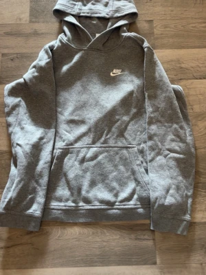 Grå hoodie från Nike med logga - Säljer en klassisk grå hoodie från Nike med vit broderad logga på bröstet. Hoodien har huva och en stor magficka framtill. Tillverkad i mjukt sweatshirtmaterial som är perfekt för chill dagar. Enkel och stilren design som funkar till det mesta.