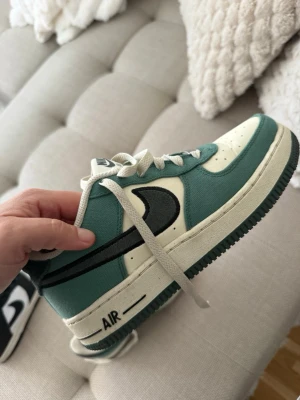 Nike Air Force 1 låga sneakers grön/vit - Nike Air Force 1 sneakers i lågt utförande med grön och vit ovandel i textil och läder. Klassisk svart swoosh på sidan och vit mellansula med AIR-detalj. Yttersulan är mörkgrön och har det ikoniska cirkelmönstret. Skorna har vita skosnören.