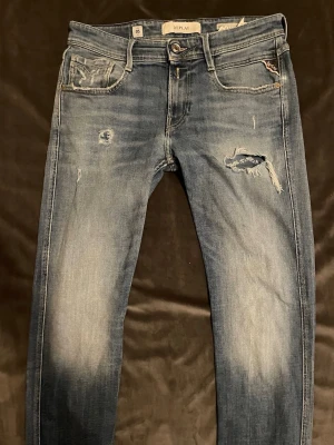 Replay jeans  - Säljer ett par blå jeans från Replay med slitningar och hål på framsidan för en cool, avslappnad look. Jeansen har fem fickor, normal midja och smal passform. Klassisk denim med ljusa tvättade partier och detaljerade sömmar.