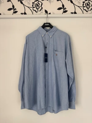 Ljusblå skjorta från Polo Ralph Lauren - Ljusblå skjorta från Polo Ralph Lauren med button-down krage och bröstficka med broderad logga. ”The big shirt” lite större i modellen. Aldrig använd och taggar kvar. Nypris ca 1500:-