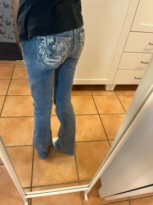 Blå bootcut jeans från miss me - Säljer ett par blå bootcut jeans med snygga broderade detaljer och nitar på bakfickorna. Jeansen har låg midja, kontrastsömmar och klassisk femficksmodell. Perfekta för dig som vill ha ett par jeans med lite extra attityd!