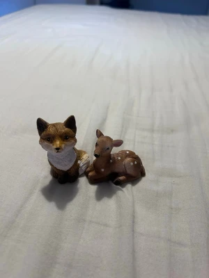 Djurfigurer räv och rådjur - Två små djurfigurer i plast, en räv och ett liggande rådjur. Räven är brun med vit mage och svans, och rådjuret är brunt med vita prickar på ryggen. Perfekta som dekoration eller leksak till samlingen.