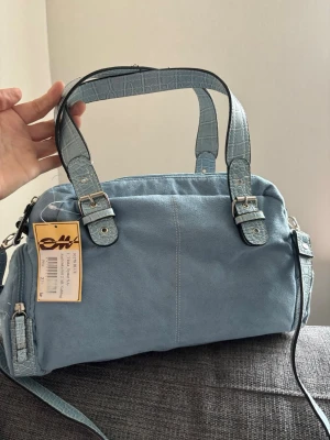 Ljusblå handväska med crocomönster - Trendig ljusblå handväska i mockaimitation med detaljer och handtag i crocomönstrat konstläder. Väskan har silverfärgade spännen och dragkedjor samt en avtagbar axelrem. Perfekt accessoar för att lyfta din outfit. Mått 25x20x11 cm 