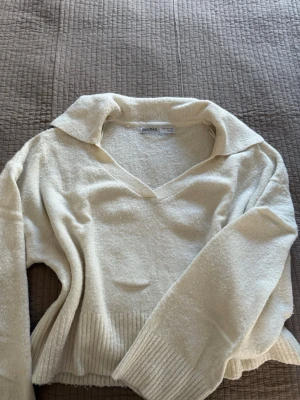 vit stickad tröja från Bershka - vit stickad tröja från Bershka med v-ringning och bred krage. Tröjan har långa ärmar och ribbstickade muddar vid ärmslut och nederkant. Perfekt för lager-på-lager och en stilren look.