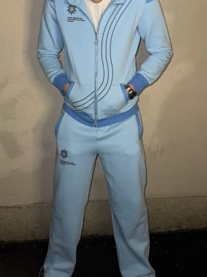 Ceyst  - Snyggt loungewear-set i ljusblått med matchande byxor och zip-hoodie. Setet har mörkare blå detaljer vid muddar och midja samt svarta linjer på jackan. Tryckt logga på både jacka och byxa. Perfekt för chill dagar hemma,eller gymbyxor 