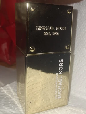 Michael Kors 24K Brilliant Gold parfym - Lyxig parfymflaska i metalliskt guld med fyrkantig form från Michael Kors. Flaskan har elegant design med texten '24K Brilliant Gold' och märkets logga ingraverad. Perfekt för dig som gillar exklusiva dofter och snygg inredningsdetalj.mer än hälften kvar 