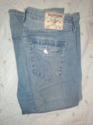 Ljusblå True Religion jeans - Säljer ett par ljusblå jeans från True Religion med klassiska kontrastsömmar och stora fickor bak med lock och silverfärgade knappar. Jeansen har en avslappnad passform och är tillverkade i denim. Perfekta för en cool och avslappnad stil. (Mid waist) pris går att diskuteras, storlek w28