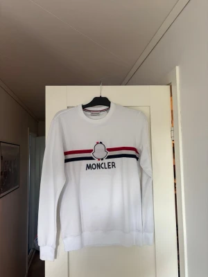 Vit Moncler sweatshirt  - Snygg vit sweatshirt från Moncler med broderad logga och text på bröstet. Tröjan har röda och marinblå ränder över bröstet och ribbade muddar vid ärmslut och nederkant. Perfekt för en clean look. (Säljes för köp i fel storlek)