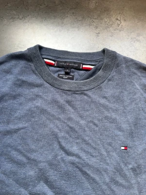 Blå stickad tröja Tommy Hilfiger - Mysig blå stickad tröja från Tommy Hilfiger i ekologisk bomull. Tröjan har rund halsringning, långa ärmar och en liten broderad logga på bröstet. Perfekt för lager på lager och enkel att matcha med jeans eller chinos. Se fläcken på bild 3
