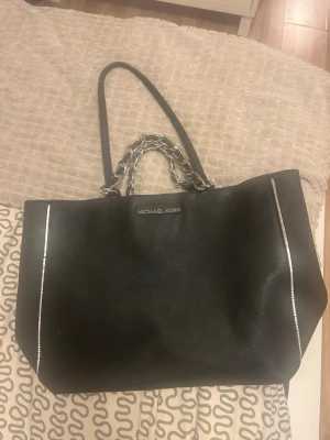 Svart Michael Kors handväska - Snygg svart handväska från Michael Kors med silverfärgade kedjedetaljer på handtagen och diskreta vita linjer längs kanterna. Väskan är rymlig och har en klassisk rektangulär form, perfekt för att bära med sig allt man behöver.