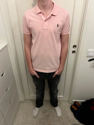 Ljusrosa pikétröja från Polo Ralph Lauren - Säljer en ljusrosa pikétröja från Polo Ralph Lauren med klassisk krage och knappar framtill. Tröjan har korta ärmar och den ikoniska loggan broderad på bröstet. Perfekt för en clean och stilren look.