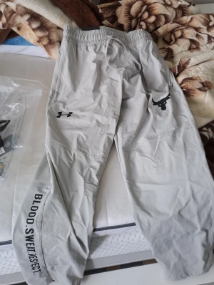 Grå Under Armour  - Säljer ett par grå mjukisbyxor från Under Armour med trycket 'Blood. Sweat. Respect.' på benet och Project Rock-loggan framtill. Byxorna har resår i midjan och muddar vid bensluten. Perfekta för chill.