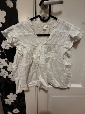 Vit blus med volangärm och v-ringning - Söt vit blus med korta volangärmar och djup v-ringning. Blusen har fina detaljer med rynk och spets längs axlar och bröst. Tillverkad i ett lätt och luftigt tyg, perfekt för sommardagar. Passar till både jeans och kjol.