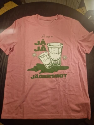 Rosa t-shirt med JÄGERSHOT-tryck - Säljer en rosa t-shirt i storlek XL med grönt tryck framtill som föreställer två shotglas och texten 'Ge mig en... JÄGERSHOT'. T-shirten har rund hals och korta ärmar. Perfekt för dig som gillar roliga motiv och festliga plagg.