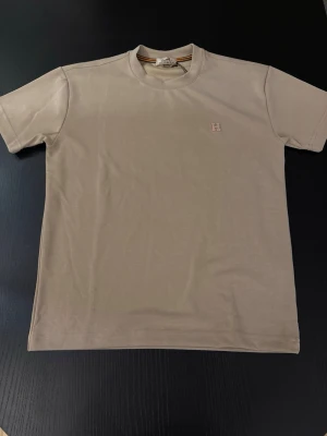 Beige kortärmad t-shirt med broderad H - Snygg beige t-shirt med korta ärmar och en liten broderad H-detalj på bröstet. Tillverkad i mjuk bomull och har en klassisk passform som passar till det mesta. Perfekt för dig som gillar stilrena och enkla plagg.