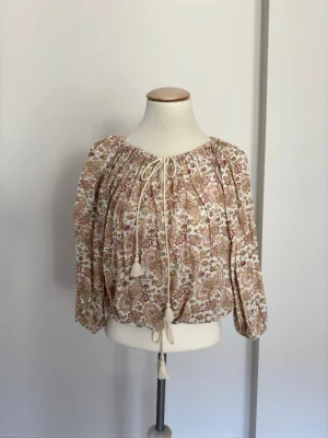 Blommig boho-blus med knytband - Romantisk blus i bohostil med blommigt mönster i beige, rosa och vinröda toner. Vida långärmar och rynkad halsringning med dekorativa knytband och tofsar. Tillverkad i ett lätt, vävt material som känns luftigt och bekvämt.