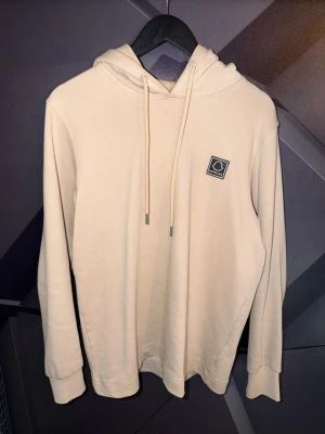 Moncler hoodie - Snygg beige hoodie från Moncler med klassisk svartvit logga på bröstet. Hoodien har huva med dragsko och ribbade muddar vid ärmslut och nederkant. Tillverkad i mjukt bomullsmaterial som känns skönt mot huden. Perfekt för en clean och stilren look.