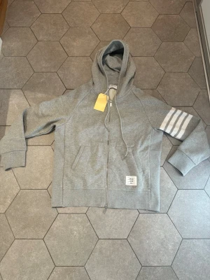 Grå hoodie med dragkedja Thome Browne  - Snygg grå Thom Browne hoodie med dragkedja framtill och huva med dragsko. På vänster ärm finns fyra vita ränder som ger en sportig look. Fickor på sidorna och en liten vit etikett nedtill. Perfekt för en avslappnad stil.