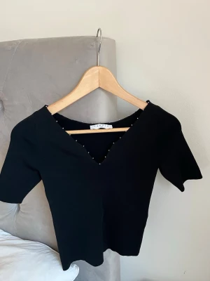 Svart topp - Snygg svart topp från Sandro med v-ringning och små silverfärgade pärlor längs halsringningen. Kortärmad och figurnära modell som passar perfekt till jeans eller kjol. Stilren och enkel design som känns lyxig. Självklart äkta!❤️