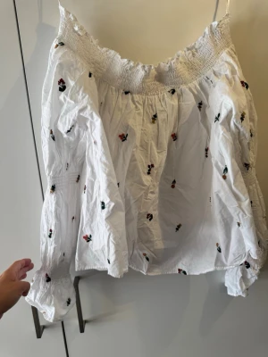 Vit blus med broderade blommor - Söt vit blus med broderade små blommor i olika färger. Blusen har långa ärmar med smock vid axlar och ärmslut samt en offshoulder-design. Perfekt för en somrig och romantisk look.