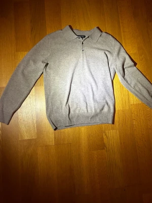 Ljusgrå stickad tröja med krage - En stilren ljusgrå/beige Grish stickad tröja med klassisk krage och knäppning framtill. Tröjan har långa ärmar och ribbade muddar vid ärmslut och nederkant. Perfekt för dig som gillar en enkel men snygg look.
