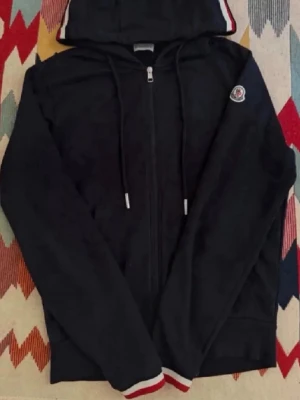 Marinblå hoodie med dragkedja - Svart hoodie med dragkedja framtill, huva med dragsko. Ribbade muddar med röd och vit detalj. Perfekt för en stilren och sportig look.