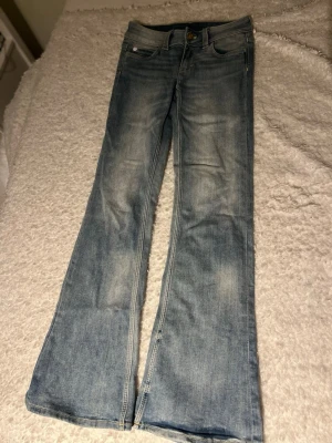 Ljusblå bootcut jeans - Säljer ett par ljusblå bootcut jeans med slitningar och klassisk femficksmodell. Jeansen har en snygg tvätt och utsvängda ben, perfekta för en avslappnad och trendig look. Materialet är denim i bomull.
