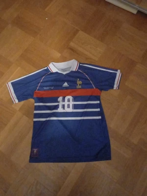 Frankrike Zidane tröja Adidas blå - Säljer en klassisk blå fotbollströja från Adidas med Frankrikes landslagsmärke och nummer 10 på bröstet. På ryggen står det Zidane och siffran 10. Tröjan har vita och röda detaljer samt krage med röd och blå rand. ALDRIG ANVÄND.