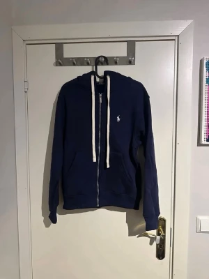 Marinblå hoodie från Polo Ralph Lauren - Snygg marinblå hoodie med dragkedja från Polo Ralph Lauren. Klassisk design med vit logga på bröstet, vita snören i huvan och två fickor framtill. Tillverkad i mjukt bomullsmaterial som är perfekt för chill dagar.