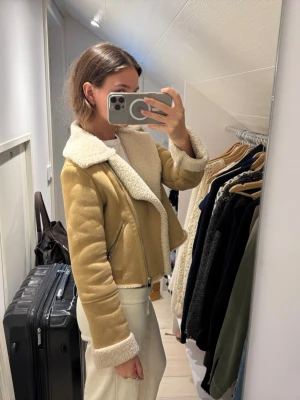 Beige teddyjacka med krage - Säljer en beige teddyjacka med vit, fluffig fuskpäls på krage, ärmslut och insida. Jackan har dragkedja framtill och två snedställda fickor med dragkedja. Kortare modell som ger en trendig look och passar perfekt till höst och vinter.