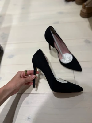 Svarta klassiska pumps i mocka - Snygga svarta pumps med spetsig tå och hög klack. Skorna är tillverkade i mocka och har en elegant siluett som passar perfekt till både jeans och klänning. Insidan är ljusrosa och klacken är smal för en stilren look.