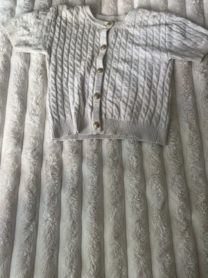 vit stickad kofta - Kort, vit kabelstickad kofta med korta ärmar och metall knappar framtill. Koftan har ribbade muddar vid ärmslut och nederkant, och ett klassiskt mönster som ger en trendig look. Perfekt att slänga över en topp för extra stil.
