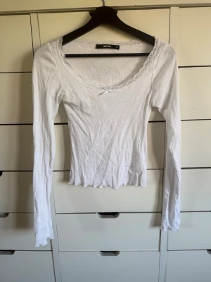 Vit långärmad topp från BikBok - Söt vit långärmad topp från BikBok med ribbad struktur, spetskant vid ringningen och en liten rosett framtill. Perfekt för dig som gillar en feminin och enkel stil. Tunn och mjuk i materialet.