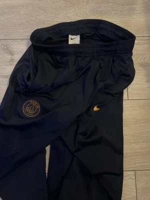 Svarta PSG träningsbyxor från Nike - Säljer ett par svarta träningsbyxor från Nike med PSG-logga på ena benet och gul Nike swoosh på andra. Byxorna har resår i midjan och smal passform, perfekta för fotboll eller träning.